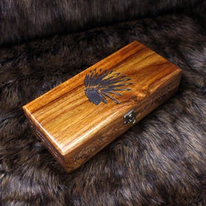 May include: A rectangular wooden box with a dark finish, featuring a carved Native American headdress design on the lid. The box has a metal clasp and appears to be made of a rich, warm-toned wood. The box is resting on a brown, furry surface.