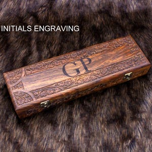 May include: A rectangular wooden box with intricate floral carvings and the initials "GP" engraved on the lid. The box is a rich brown color and has a clasp closure. The text "INITIALS ENGRAVING" is above the box.