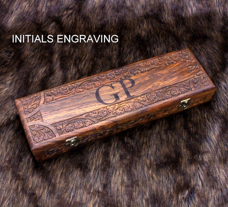 May include: A rectangular wooden box with intricate floral carvings and the initials "GP" engraved on the lid. The box is a rich brown color and has a clasp closure. The text "INITIALS ENGRAVING" is above the box.