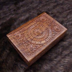 May include: A rectangular wooden box with detailed floral carvings. The box is a warm brown colour and features a detailed design of flowers and swirling patterns. The lid is adorned with a central circular motif and is surrounded by more floral elements.