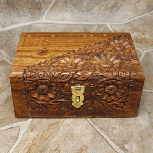 Puede incluir: Una caja de madera rectangular con intrincadas tallas florales y un cierre de lat&oacute;n. La caja es de color marr&oacute;n c&aacute;lido, con tallas detalladas de flores y hojas. La parte superior tiene dise&ntilde;os incrustados de estrellas y puntos de lat&oacute;n.