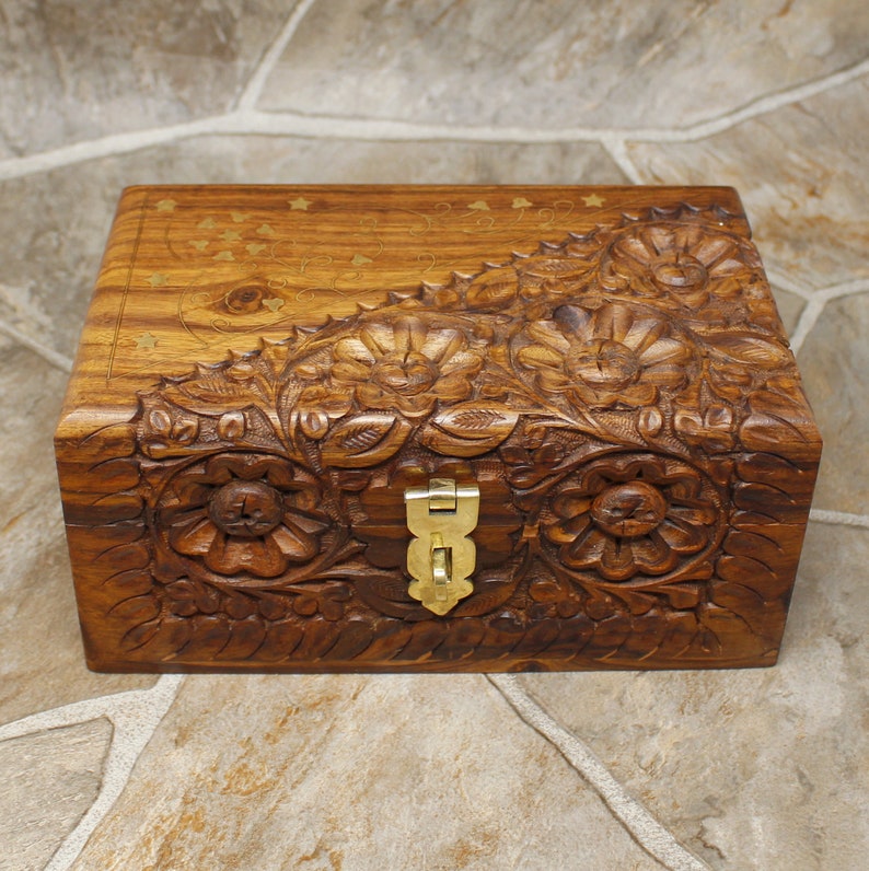 Puede incluir: Una caja de madera rectangular con intrincadas tallas florales y un cierre de lat&oacute;n. La caja es de color marr&oacute;n c&aacute;lido, con tallas detalladas de flores y hojas. La parte superior tiene dise&ntilde;os incrustados de estrellas y puntos de lat&oacute;n.