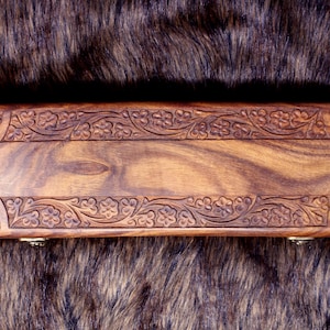 May include: A rectangular wooden box with intricate floral carvings along the edges. The box is a warm brown color, with a visible wood grain pattern. The box is resting on a dark, textured surface.