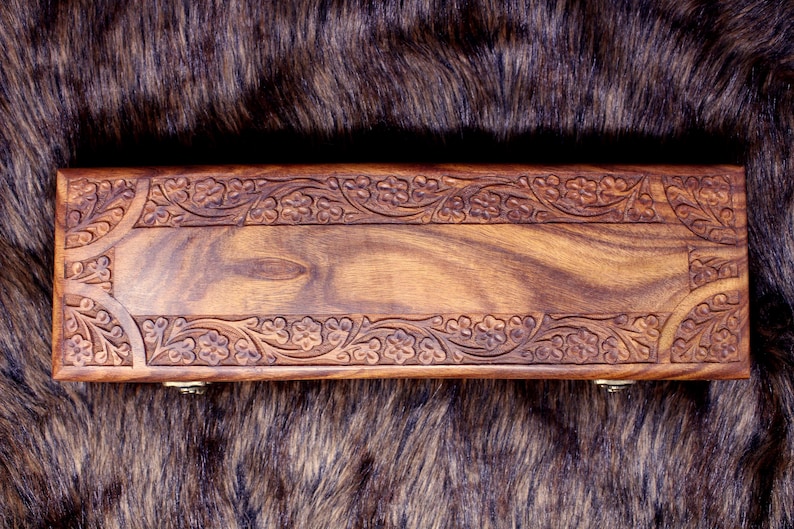May include: A rectangular wooden box with intricate floral carvings along the edges. The box is a warm brown color, with a visible wood grain pattern. The box is resting on a dark, textured surface.