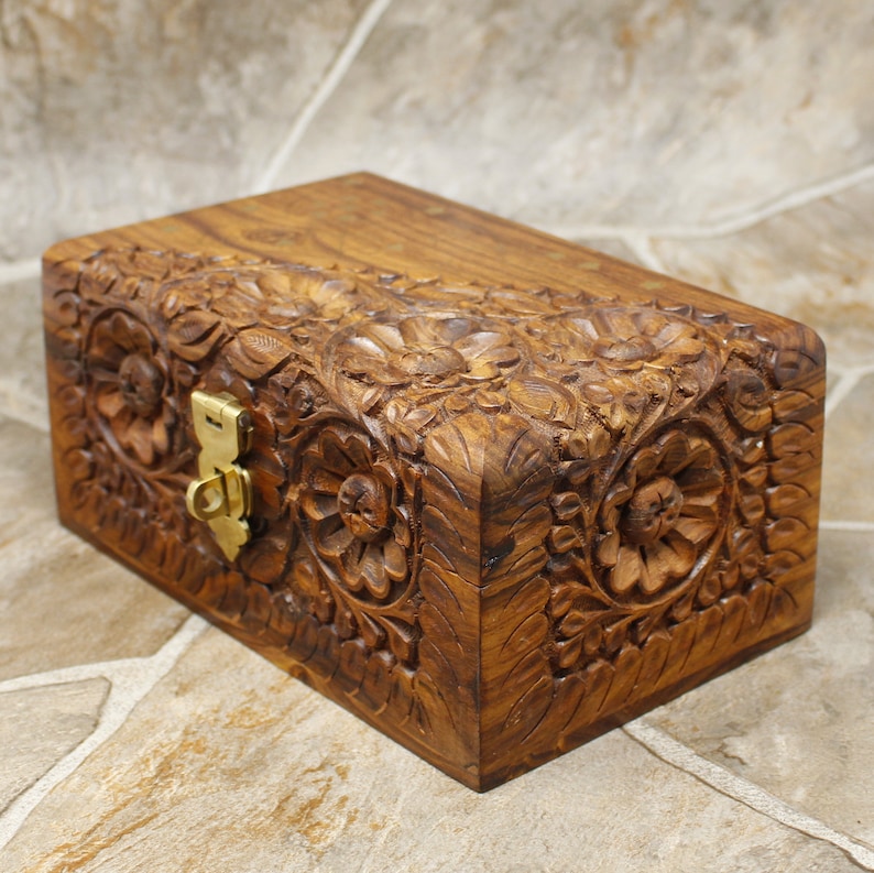 Puede incluir: Una caja rectangular de madera con intrincadas tallas florales y un cierre de lat&oacute;n. La caja es de color marr&oacute;n c&aacute;lido y presenta detalladas tallas en relieve de flores y hojas en su superficie. Mide aproximadamente 20 cm de largo, 13 cm de ancho y 10 cm de alto.