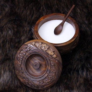 May include: A wooden salt cellar with a carved floral design on the lid. The open container is filled with white salt, and a small wooden spoon rests inside. The dark brown wood contrasts with the white salt, creating a rustic aesthetic.