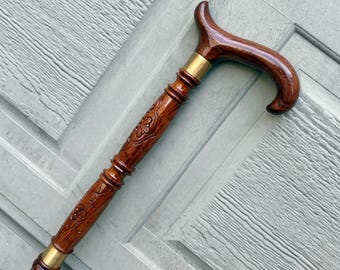 May include: A wooden cane with a curved handle and a detailed floral pattern carved into the shaft. The cane features brass accents and a rich, dark brown finish, offering a classic and elegant look.
