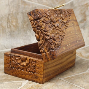 Puede incluir: Un joyero de madera con intrincadas tallas florales. La caja est&aacute; abierta, revelando el interior, y presenta un dise&ntilde;o floral tallado en la tapa y el frente. La madera tiene un tono marr&oacute;n c&aacute;lido, sobre un fondo neutro.