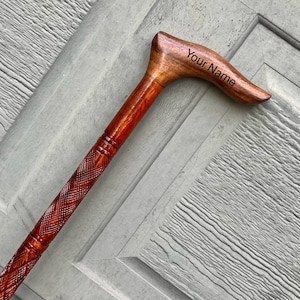 May include: A wooden walking stick with a carved handle. The handle is curved and has the words "Your Name" engraved on it. The stick is brown and has a reddish-brown stain.