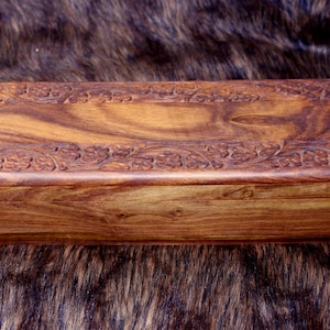 May include: A rectangular wooden box with a hinged lid. The box is made of a warm brown wood with a natural grain pattern. The lid and sides are adorned with carved floral designs. The box is approximately 10 inches long.