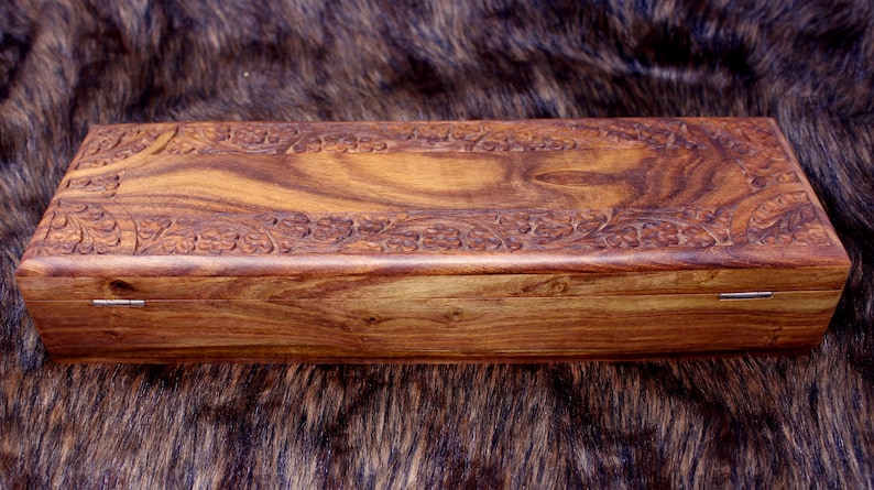 May include: A rectangular wooden box with a hinged lid. The box is made of a warm brown wood with a natural grain pattern. The lid and sides are adorned with carved floral designs. The box is approximately 10 inches long.