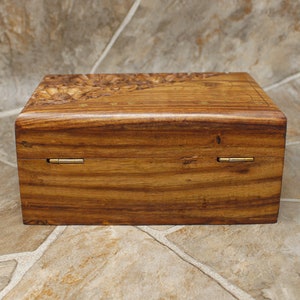 Puede incluir: Caja rectangular de madera con un dise&ntilde;o floral tallado en la tapa. La caja es de madera de tono c&aacute;lido, con vetas visibles y bisagras de lat&oacute;n. El dise&ntilde;o general es simple y elegante.