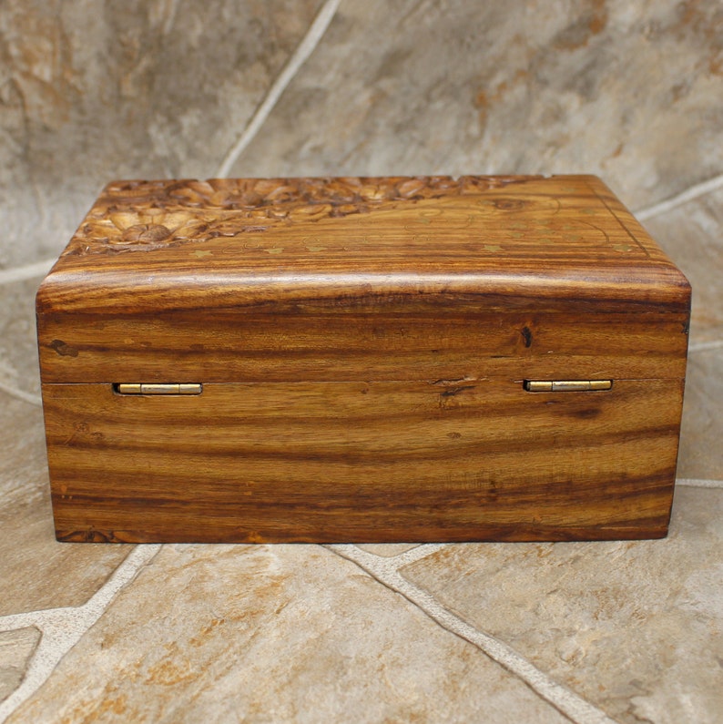 Puede incluir: Caja rectangular de madera con un dise&ntilde;o floral tallado en la tapa. La caja es de madera de tono c&aacute;lido, con vetas visibles y bisagras de lat&oacute;n. El dise&ntilde;o general es simple y elegante.