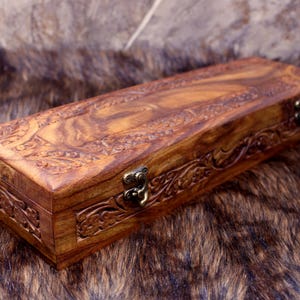 May include: A rectangular wooden box with intricate carved floral designs. The box is a warm brown color with a hinged lid and a metal clasp. The box is resting on a brown and tan fur-like surface.