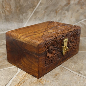 Puede incluir: Una caja rectangular de madera con intrincadas tallas florales y un cierre de lat&oacute;n. La caja es de madera marr&oacute;n oscuro y tiene un acabado liso y pulido. El dise&ntilde;o floral est&aacute; tallado en la parte superior y frontal.