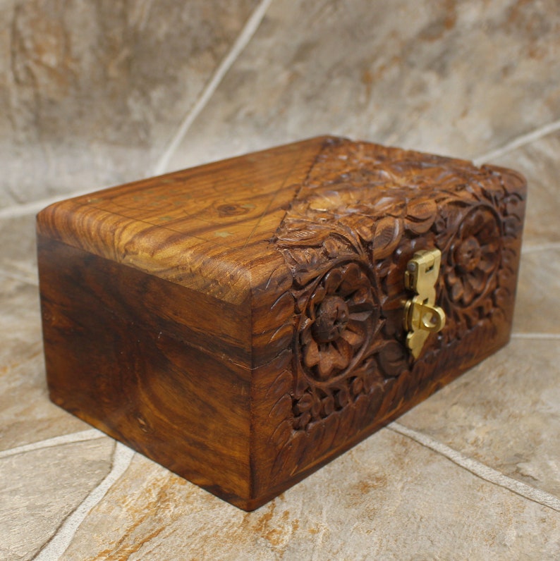 Puede incluir: Una caja rectangular de madera con intrincadas tallas florales y un cierre de lat&oacute;n. La caja es de madera marr&oacute;n oscuro y tiene un acabado liso y pulido. El dise&ntilde;o floral est&aacute; tallado en la parte superior y frontal.
