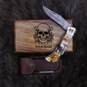 May include: A folding knife with a patterned blade and a handle with a bone-like appearance. The knife rests on a wooden box with a skull and rose design and the text "YOUR NAME". A leather sheath is also present.