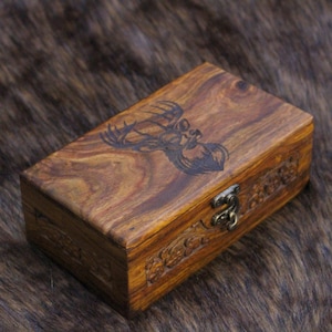 Peut inclure: Une boîte en bois rectangulaire avec un motif de cerf gravé sur le couvercle. La boîte a un fermoir en laiton et des motifs floraux sculptés sur les bords. Le grain du bois est visible et la boîte est de couleur marron chaud.