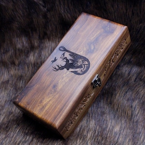 May include: A rectangular wooden box with a dark brown finish. The lid features a carved design of a deer, trees, and a fish hook. The box has a metal clasp and decorative carvings along the sides, suggesting a rustic or outdoors theme.