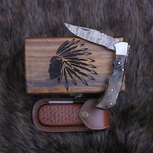 May include: A Damascus steel folding knife with a wooden handle and a leather sheath. The knife is next to a wooden box with an engraved Native American headdress design. The knife blade is open, revealing its intricate pattern.