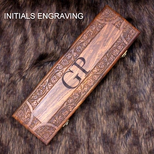 May include: A rectangular wooden box with floral carvings and the initials "GP" engraved on the lid. The box is a warm brown color with gold-colored hinges. The text "INITIALS ENGRAVING" is at the top.