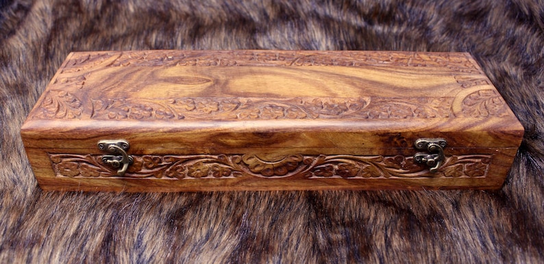 May include: A rectangular wooden box with intricate floral carvings on the lid and sides. The box is a warm brown color with a visible wood grain pattern. It features a metal clasp closure and is set against a textured background.
