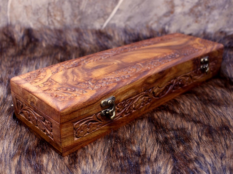 May include: A rectangular wooden box with intricate carved floral designs. The box is a warm brown color with a hinged lid and a metal clasp. The box is resting on a brown and tan fur-like surface.