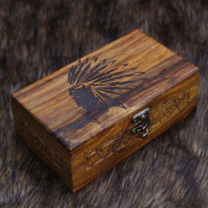 May include: A rectangular wooden box with a hinged lid and metal clasp. The box is a warm brown color with a carved Native American headdress design on the lid. The box has carved details around the edges.