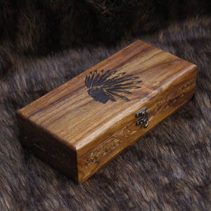 May include: A rectangular wooden box with a dark finish. The lid features a black engraved Native American headdress design. The box has a metal clasp and carved floral details along the sides. The box is on a brown, furry surface.