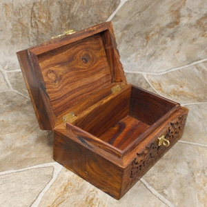 Puede incluir: Caja de madera marr&oacute;n oscuro con tapa con bisagras abierta. La caja presenta intrincadas tallas florales y un cierre de lat&oacute;n. El interior est&aacute; vac&iacute;o, revelando la veta de la madera. Caja de almacenamiento, aproximadamente 15 cm de largo.