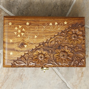Puede incluir: Caja de madera con intrincadas tallas florales e incrustaciones de lat&oacute;n. La tapa presenta un dise&ntilde;o de coraz&oacute;n y estrellas, con un cierre de lat&oacute;n. La madera tiene un tono marr&oacute;n c&aacute;lido y la caja parece hecha a mano.