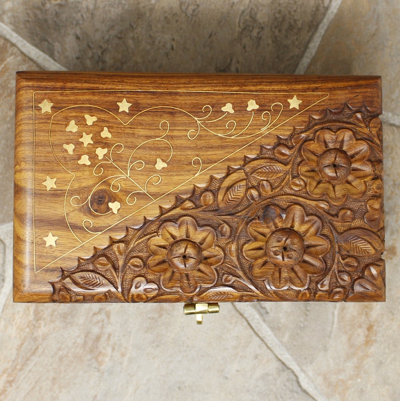 Puede incluir: Caja de madera con intrincadas tallas florales e incrustaciones de lat&oacute;n. La tapa presenta un dise&ntilde;o de coraz&oacute;n y estrellas, con un cierre de lat&oacute;n. La madera tiene un tono marr&oacute;n c&aacute;lido y la caja parece hecha a mano.
