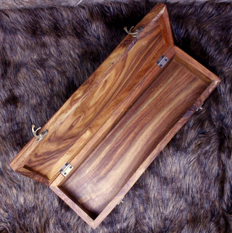 May include: Open wooden box with a hinged lid. The box is made of a light brown wood with a visible grain pattern. The interior is empty. The box is on a brown and black fur-like surface. The box has brass-colored clasps.