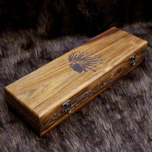 May include: A rectangular wooden box with a Native American headdress design etched on the lid. The box is made of light brown wood with a decorative carved border and metal clasps.