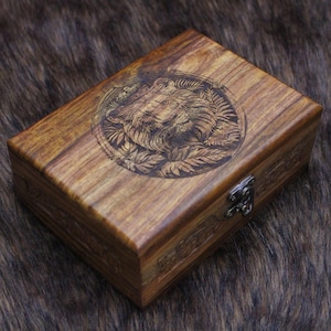 May include: A rectangular wooden box with a detailed engraved lion's head surrounded by leaves on the lid. The box is a warm brown color with a metal clasp and intricate carvings along the edges. The box is likely used for storage.