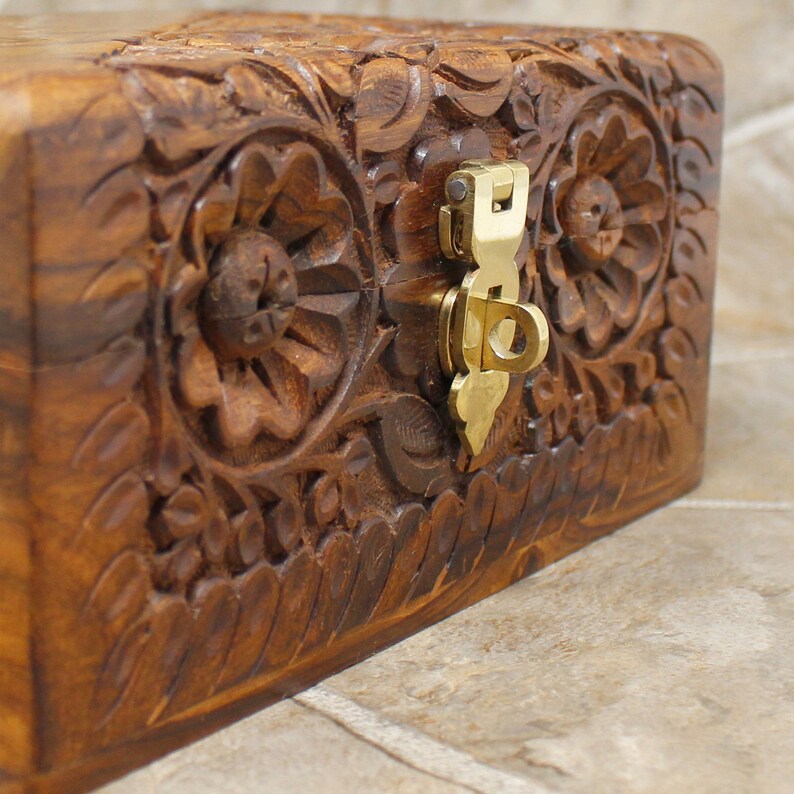 Puede incluir: Una caja de madera rectangular con intrincadas tallas florales y un cierre de lat&oacute;n. La caja es de color marr&oacute;n c&aacute;lido, y las tallas son detalladas y texturizadas. El cierre es de color dorado brillante.
