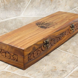 Peut inclure: Une boîte rectangulaire en bois avec un couvercle à charnière. Le bois est brun rougeâtre avec un grain visible. Le couvercle présente un motif gravé de coiffe amérindienne. Les côtés sont ornés de motifs floraux sculptés et fixés par des fermoirs couleur laiton.