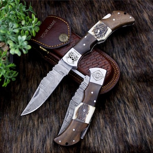 May include: Two folding knives with wooden handles and patterned blades are shown. One knife has a skull design and the name "MICHAEL". The other knife has the text "YOUR NAME". A brown leather sheath is visible.