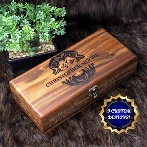 May include: A wooden box with a dark brown finish, laser-etched with a lion design and the name "CHRISTOPHER MOORE." The box has a metal clasp and is displayed on a textured, brown surface. A small plant in a black pot is in the background.