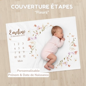 Può includere: Una coperta bianca per bambini con dettagli floreali e il testo "COUVERTURE ÉTAPES" e "Fleurs". La coperta presenta un design floreale, uno spazio per un nome e una data e i numeri 1-12. Un bambino con una tutina rosa è sdraiato sulla coperta.