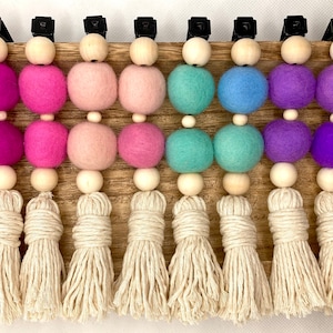 May include: A set of 10 colorful felt ball garlands with wooden beads and tassels. The garlands are in shades of pink, blue, purple, and green. Each garland has two felt balls and a wooden bead, followed by a tassel.