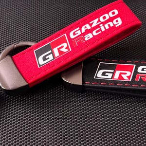 Puede incluir: Dos llaveros con el texto "GAZOO Racing" y el logotipo "GR". Uno es rojo con texto blanco, el otro es negro con texto blanco y costuras rojas. Ambos tienen un llavero de metal.
