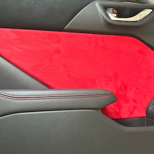 May include: Close-up of a car's interior door panel with a red suede-like material covering the armrest area. The door panel is black with red stitching.