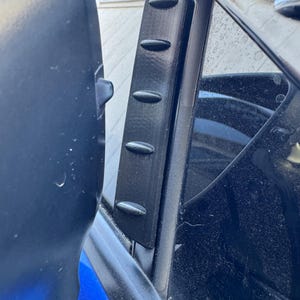 Puede incluir: Un deflector de ventana de coche negro, impreso en 3D, con un diseño aerodinámico. El deflector tiene una serie de crestas ovaladas a lo largo de su longitud. La imagen muestra el deflector instalado en una ventana de coche, con una carrocería azul visible.