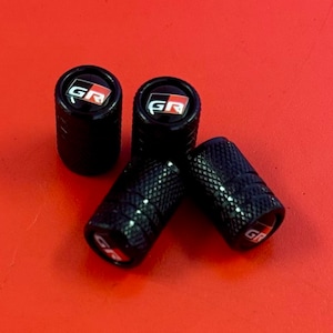 Puede incluir: Cuatro tapones de v&aacute;lvula de neum&aacute;tico negros con un logotipo "GR" rojo y blanco.