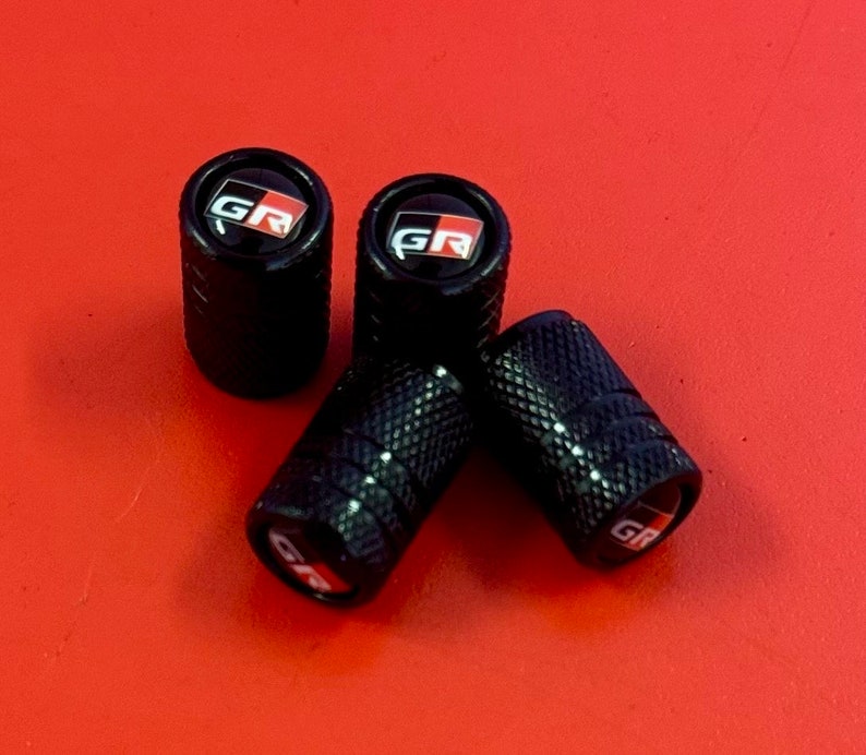 Puede incluir: Cuatro tapones de v&aacute;lvula de neum&aacute;tico negros con un logotipo "GR" rojo y blanco.
