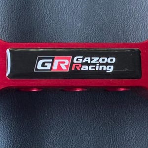 Könnte beinhalten: Ein leuchtend roter Domstrebe für Autos mit dem Text "GR Gazoo Racing" auf einer schwarzen Platte. Die Stange hat eine rechteckige Form mit mehreren Öffnungen und Befestigungslöchern, zur Leistungssteigerung.