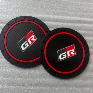 Pode incluir: Dois porta-copos redondos pretos para carro com uma borda vermelha e as letras "GR" em branco e vermelho. Os porta-copos têm uma borda externa texturizada e são projetados para caber no porta-copos de um carro.