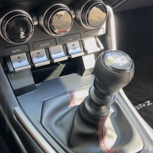 Puede incluir: Una palanca de cambios de cuero negro con costuras rojas y una base plateada. La palanca tiene una placa plateada con el texto "6 Speed 2.4L". La palanca está rodeada de un interior de coche negro y plateado con una variedad de botones y controles.