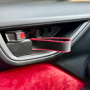 Op de afbeelding: Zwarte en rode autodeurgreep trekband. De band is bevestigd aan de deurgreep en is zwart met een rode rand. De deurgreep is zwart en heeft een rode ontgrendelingsknop. Het interieur van de auto is zwart en rood.